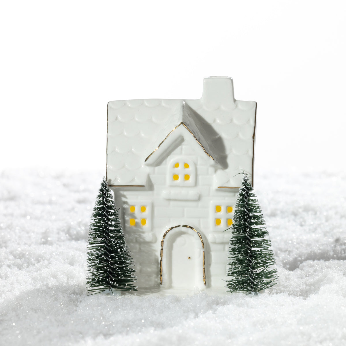 Small white ceramic brick house LED decoration featuring glowing windows, gold-trimmed details and two miniature Christmas trees on a snowy surface.