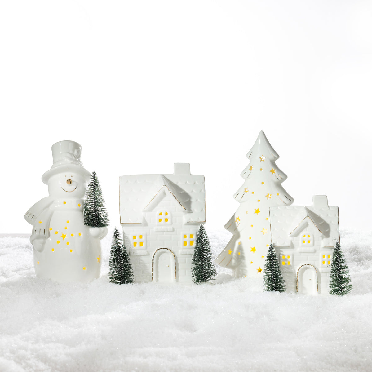 White ceramic Christmas LED decorations featuring a snowman, Christmas tree and illuminated houses with gold detailing, displayed together on a snowy surface.
