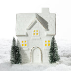 Large white ceramic Christmas brick house LED decoration featuring glowing windows, gold detailing and two miniature Christmas trees on a snowy surface.