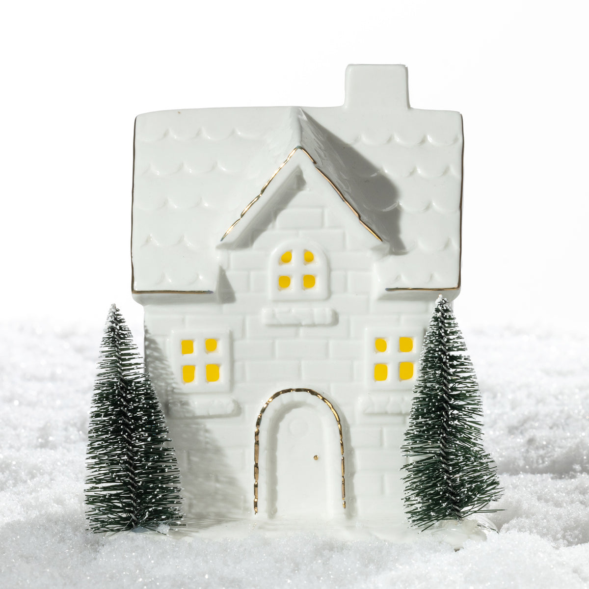 Large white ceramic Christmas brick house LED decoration featuring glowing windows, gold detailing and two miniature Christmas trees on a snowy surface.