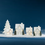 A collection of illuminated white ceramic Christmas houses and a Christmas tree with warm glowing lights, displayed on snow against a dark blue background.
