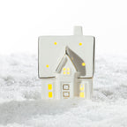White ceramic Christmas house LED decoration featuring a pitched roof with glowing dots, illuminated windows and gold-trimmed details, displayed on snow.