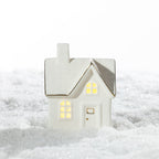 White ceramic Christmas house LED decoration featuring a brick-patterned roof, warm glowing windows and delicate gold detailing, displayed on a snowy surface.