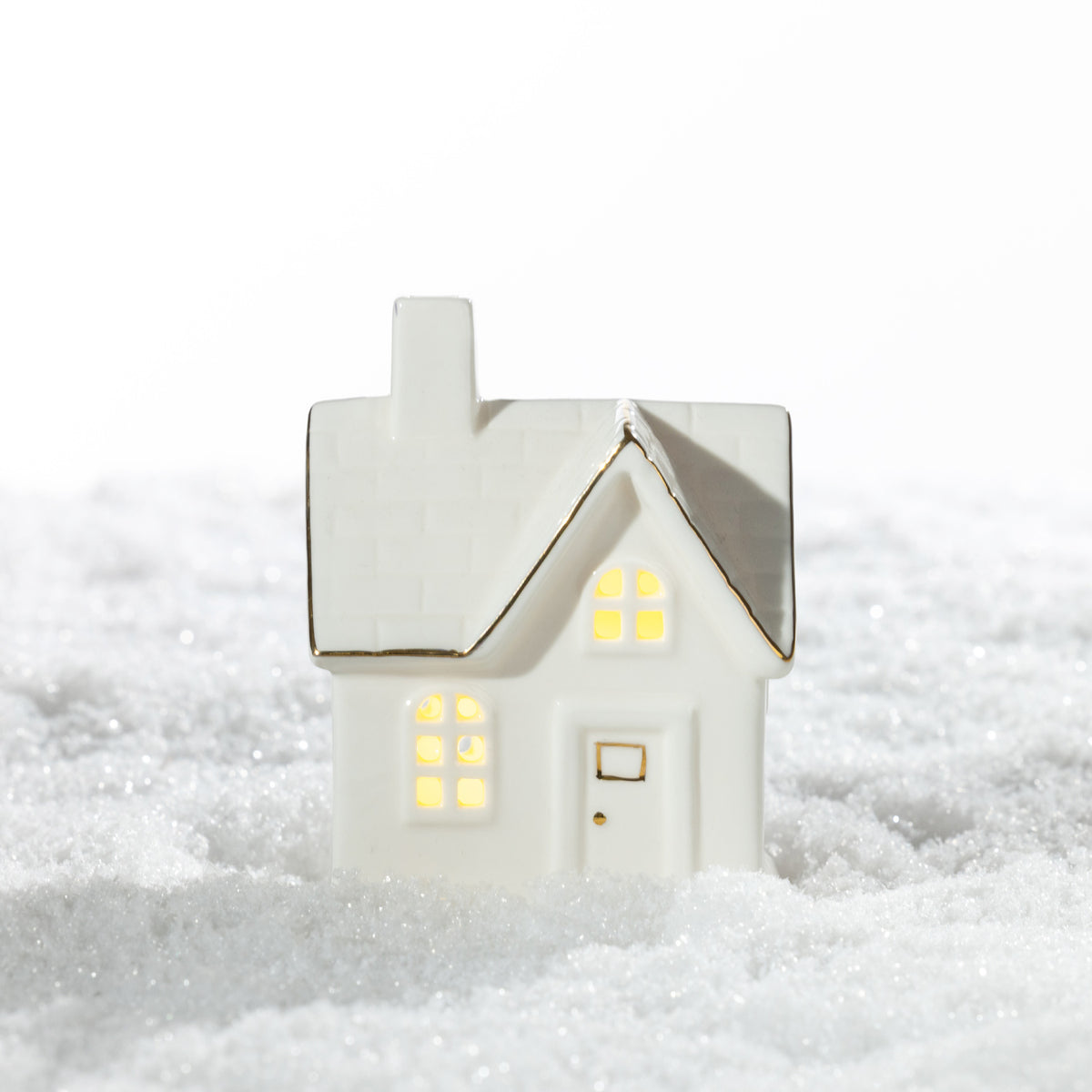 White ceramic Christmas house LED decoration featuring a brick-patterned roof, warm glowing windows and delicate gold detailing, displayed on a snowy surface.