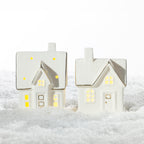 Two white ceramic Christmas houses with gold detailing and warm glowing LED windows, displayed together on a snowy surface.