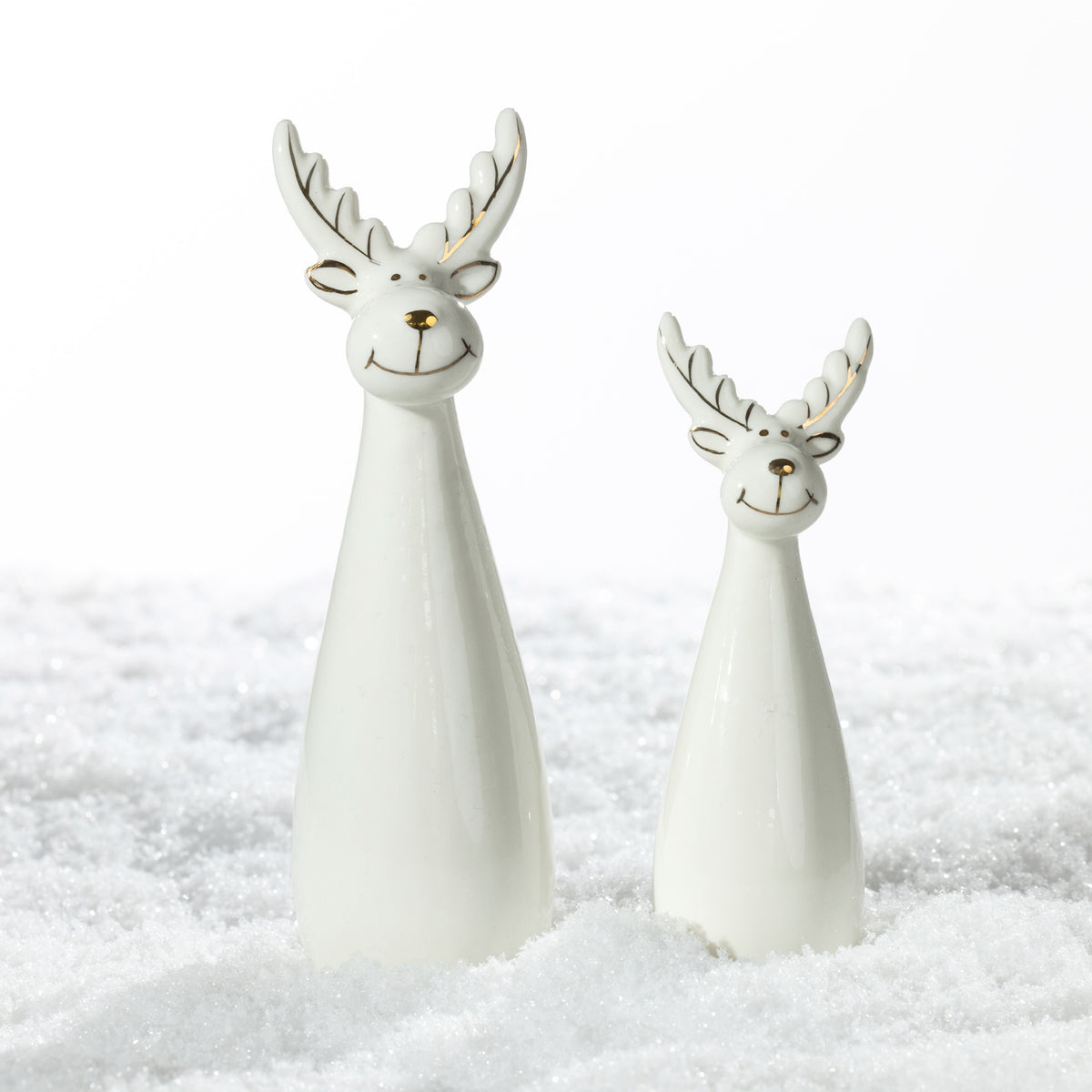 Two white ceramic Christmas reindeer decorations in different sizes, featuring smiling faces, gold-accented antlers and glossy finishes, displayed on a snowy surface.