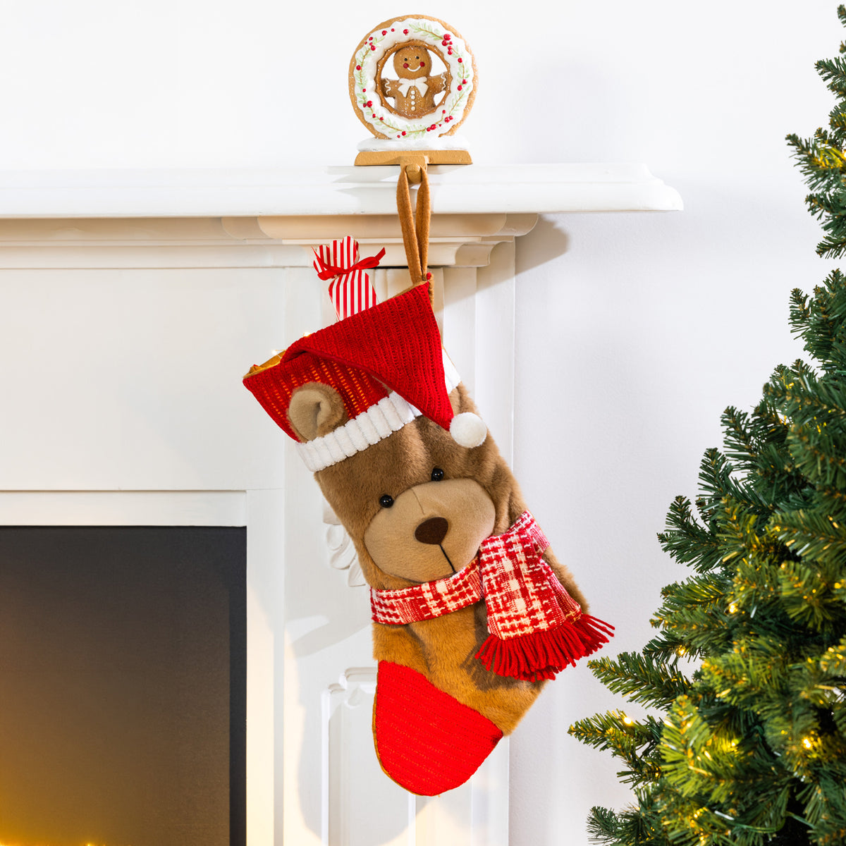 Gingerbread Christmas stocking holder featuring a smiling gingerbread man inside a festive wreath, holding a red teddy bear stocking beside a decorated Christmas tree.