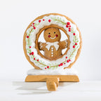 Gingerbread Christmas stocking holder featuring a smiling gingerbread man with white icing details, surrounded by a festive wreath decorated with red berries and green foliage.