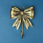 Antique gold Christmas bow wall hook featuring a textured metallic finish, flowing ribbon tails and a curved stocking hook.