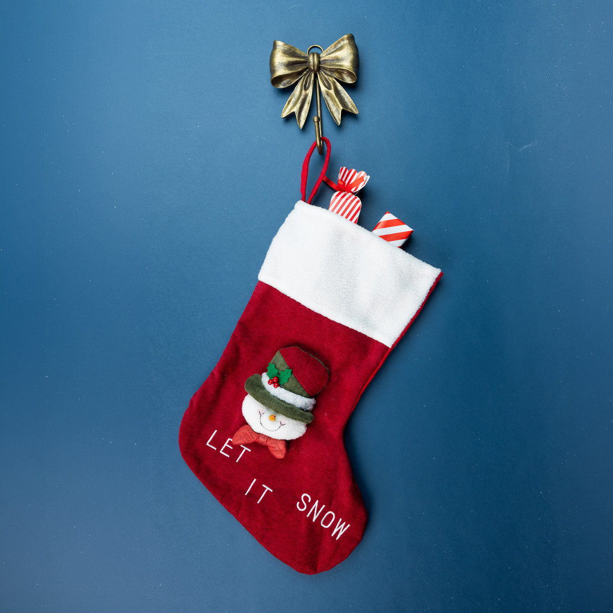 Antique gold bow wall hook holding a red Christmas stocking with a snowman design and white fluffy cuff against a blue background.