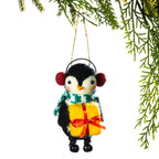 A felt penguin Christmas ornament wearing red pom-pom earmuffs, a green and white patterned scarf, and holding a yellow gift box with a red ribbon, hanging from a string against a white background with green pine branches at the top.