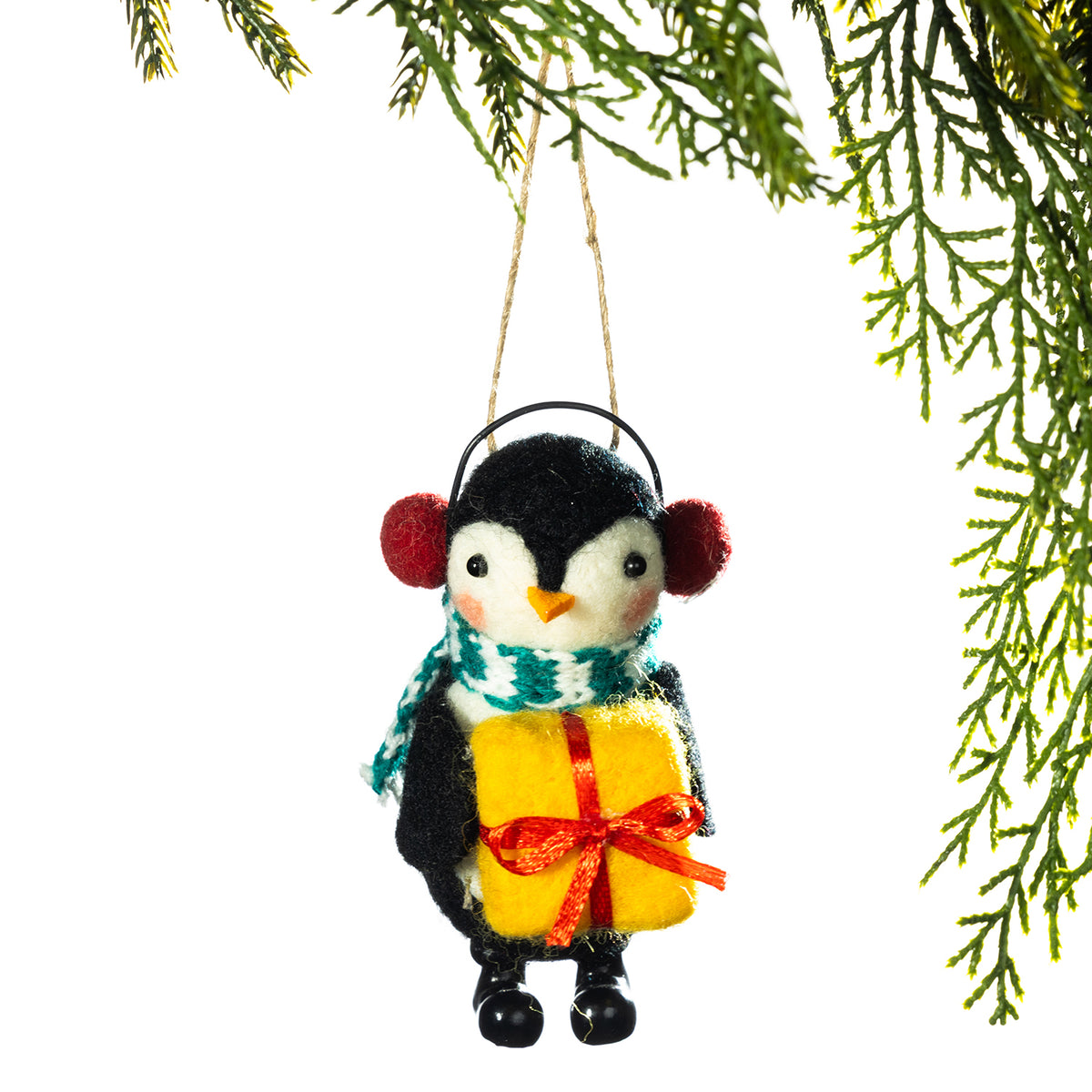 A felt penguin Christmas ornament wearing red pom-pom earmuffs, a green and white patterned scarf, and holding a yellow gift box with a red ribbon, hanging from a string against a white background with green pine branches at the top.