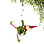 A felt Christmas elf ornament hanging from a green pine branch against a white background. The elf is green with a red hat, red-and-white striped legs, and holds a string of small lights.