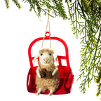 A festive Christmas ornament shaped like a fluffy cream-colored sheep sitting in a red ski lift chair, holding a small brown camera. The ornament is hanging by a rope from green pine branches against a solid white background.