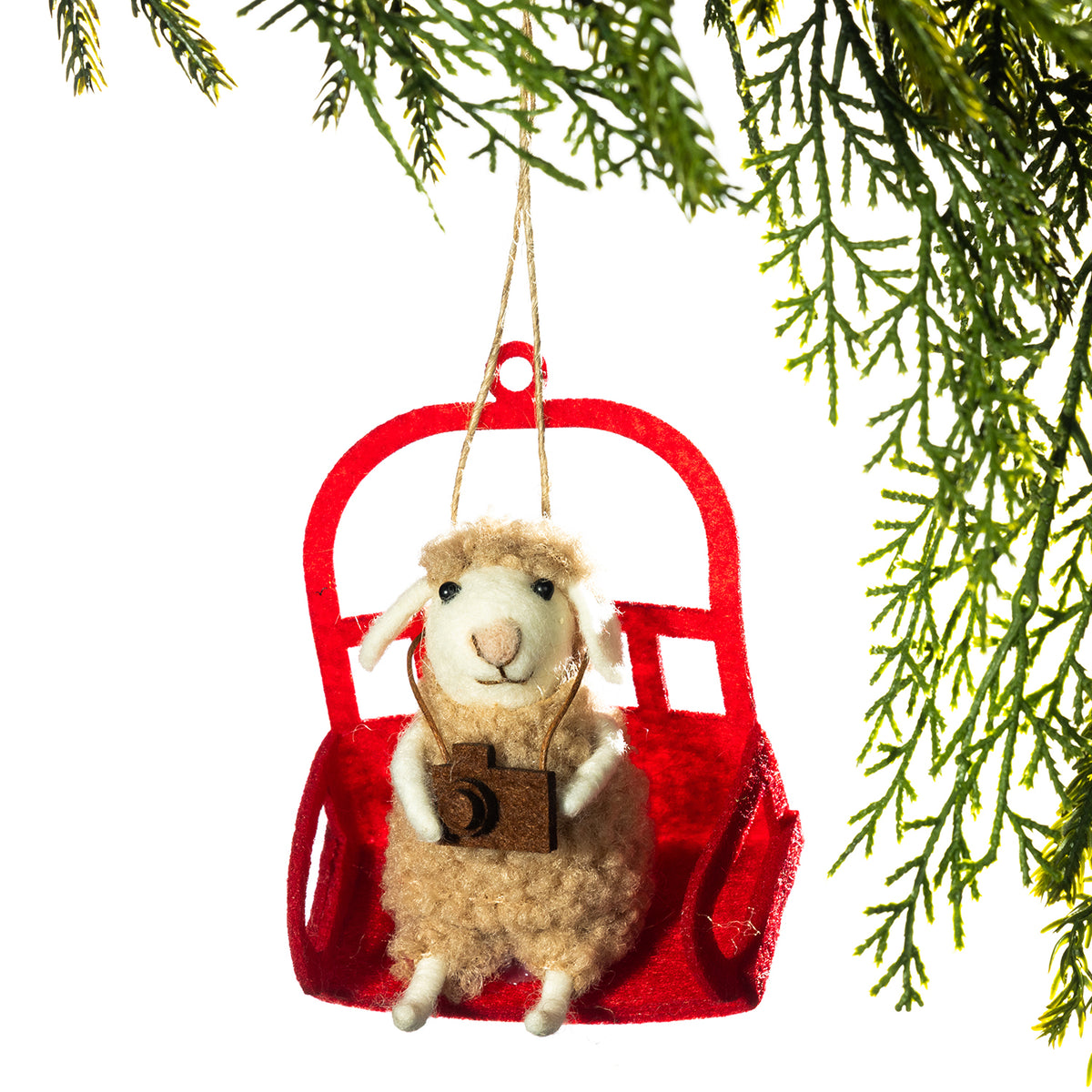 A festive Christmas ornament shaped like a fluffy cream-colored sheep sitting in a red ski lift chair, holding a small brown camera. The ornament is hanging by a rope from green pine branches against a solid white background.
