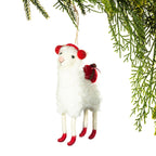 A white plush sheep Christmas ornament with red pom-pom ears, a red bow, and red hooves, hanging from a tree branch against a white background.