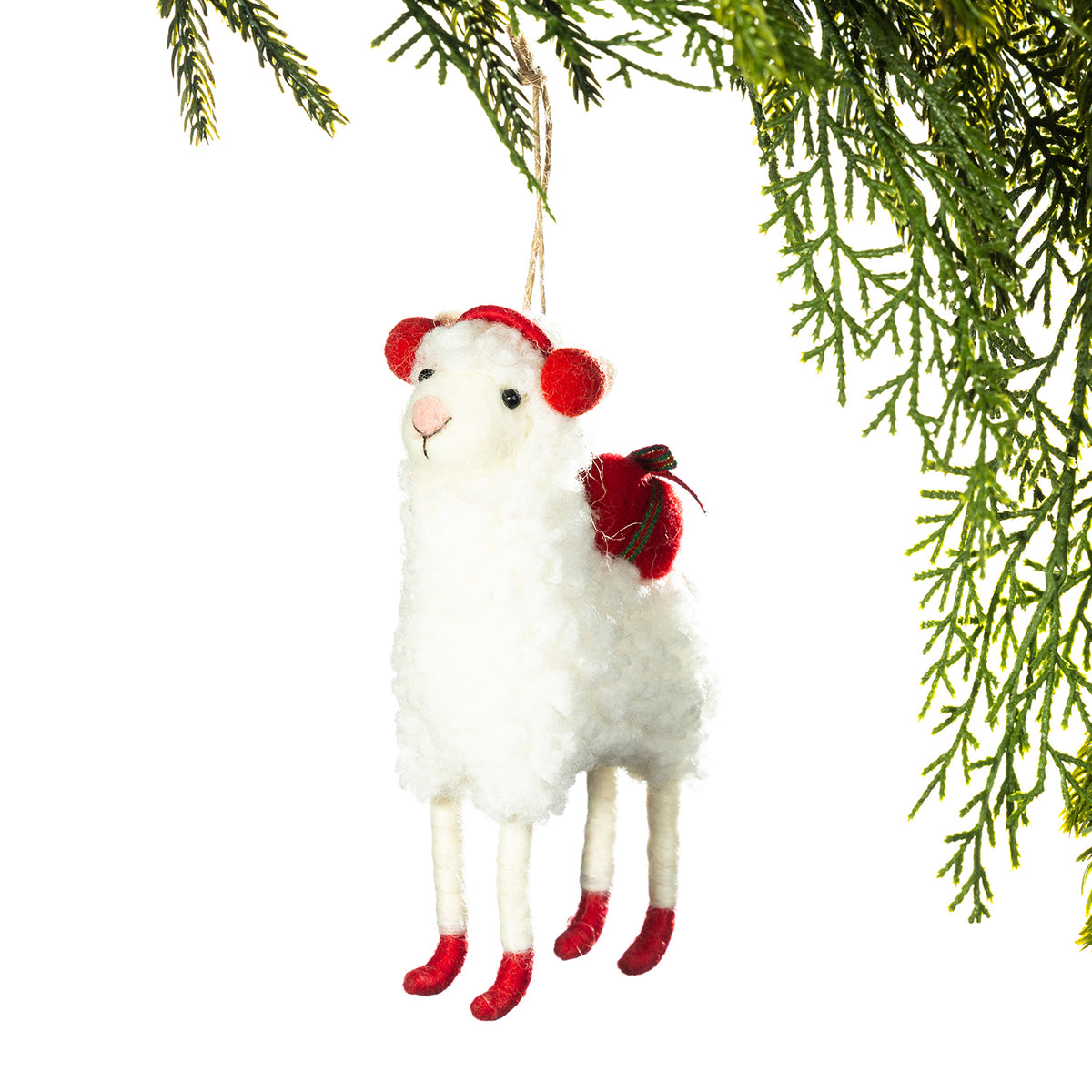 A white plush sheep Christmas ornament with red pom-pom ears, a red bow, and red hooves, hanging from a tree branch against a white background.