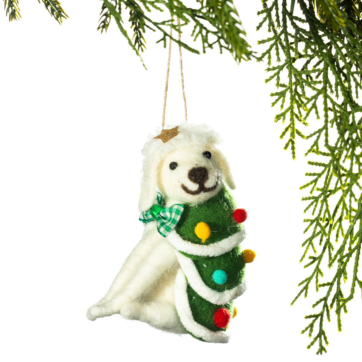 A felt Christmas ornament shaped like a white dog wrapped around a green Christmas tree with colorful pom-pom lights and a green plaid bow, hanging against a white background with evergreen branches framing the top and right edges.