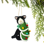 A handmade felt Christmas ornament shaped like a black and white cat wrapped in a green Christmas tree, hanging from a tree branch against a white background.