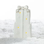 Large white ceramic stacked Christmas presents LED decoration featuring a gold-trimmed bow, embossed snowflakes and warm glowing lights on a snowy surface.