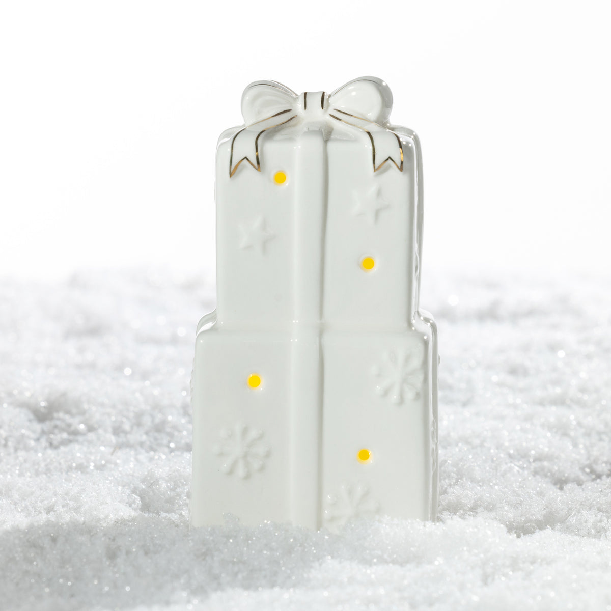 Large white ceramic stacked Christmas presents LED decoration featuring a gold-trimmed bow, embossed snowflakes and warm glowing lights on a snowy surface.