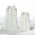 Large and small white ceramic stacked Christmas present decorations with gold-trimmed bows, embossed snowflakes and glowing LED lights, displayed on a snowy surface.