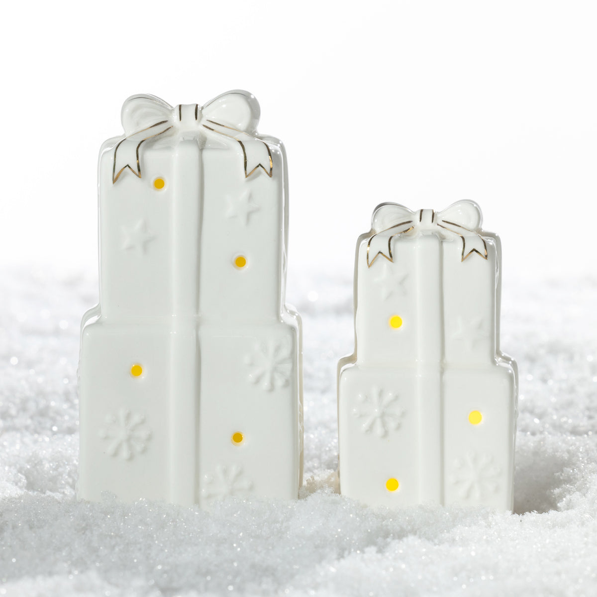 Large and small white ceramic stacked Christmas present decorations with gold-trimmed bows, embossed snowflakes and glowing LED lights, displayed on a snowy surface.
