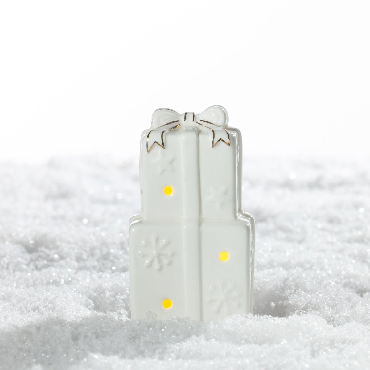 Small white ceramic stacked Christmas presents LED decoration featuring a gold-trimmed bow, embossed snowflakes and warm glowing lights on a snowy surface.