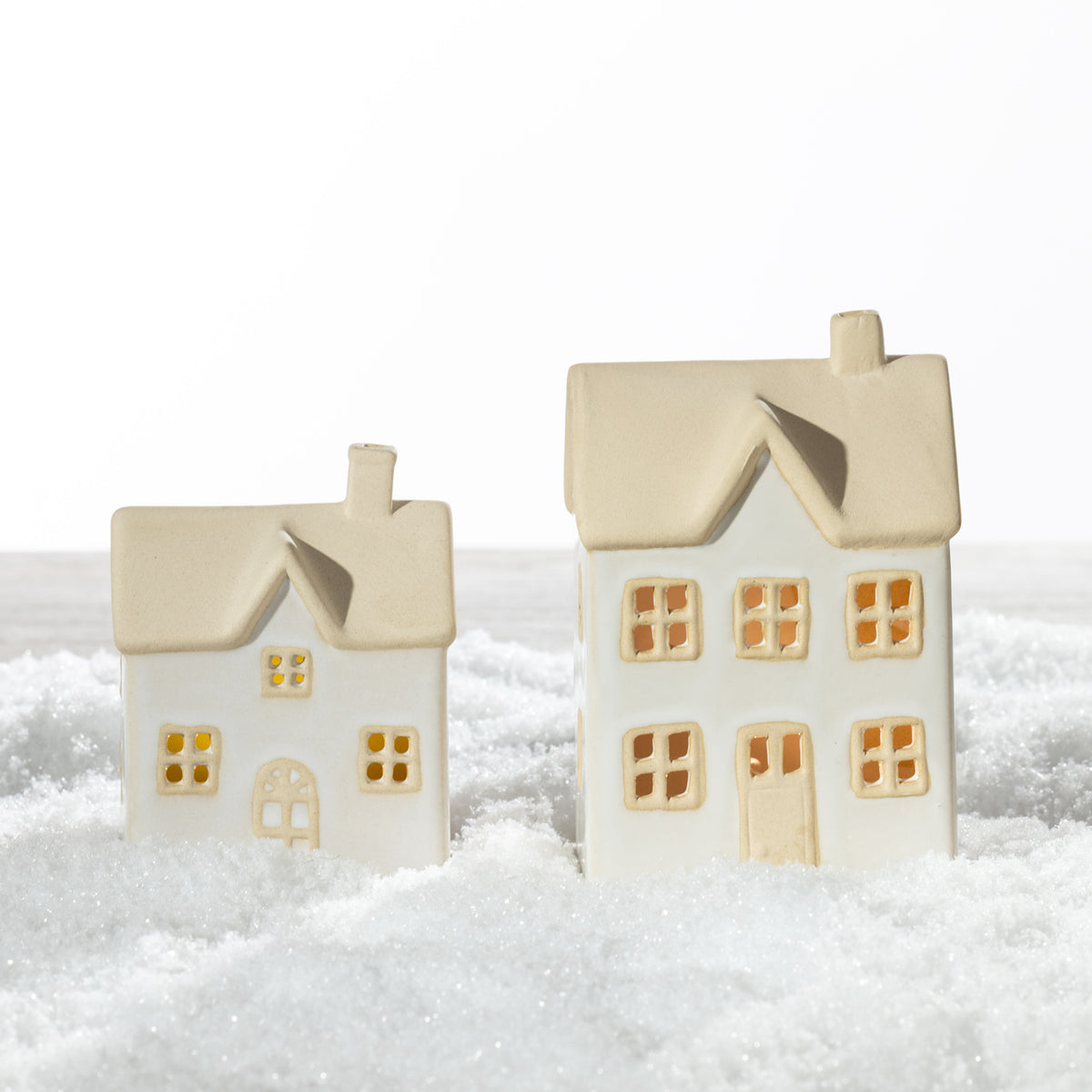 Small and large white ceramic Christmas houses with sand-coloured roofs, detailed windows and warm LED lighting, displayed together on a snowy surface.