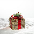 Gold rattan present box LED decoration with a red metallic bow and festive greenery, illuminated with warm lights on a snowy background.