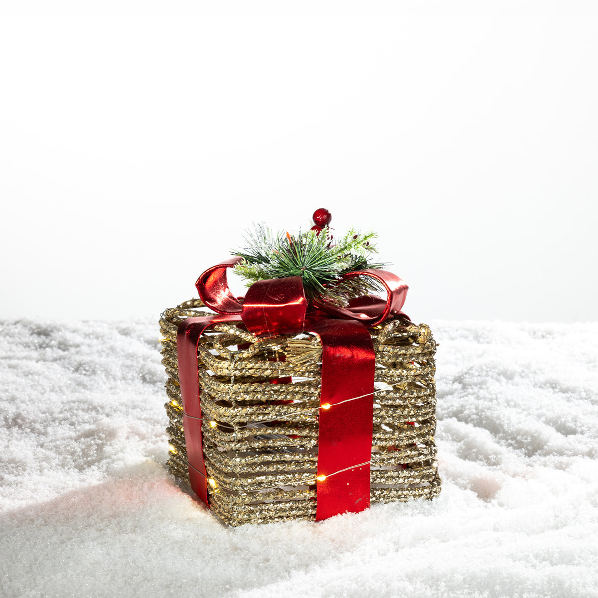 Gold rattan present box LED decoration with a red metallic bow and festive greenery, illuminated with warm lights on a snowy background.