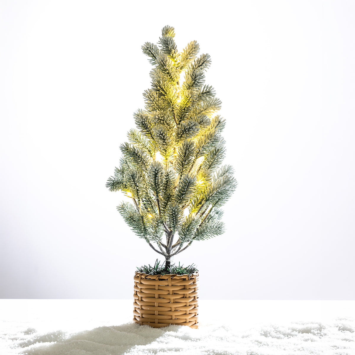 Frosted potted Christmas tree illuminated with warm LED lights, finished with a natural woven basket base on a snowy white background.