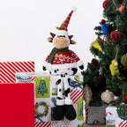 A Christmas-themed cow plush figurine wearing a red patterned sweater and a green-and-red Santa hat, posed on top of wrapped gift boxes beside a decorated Christmas tree.