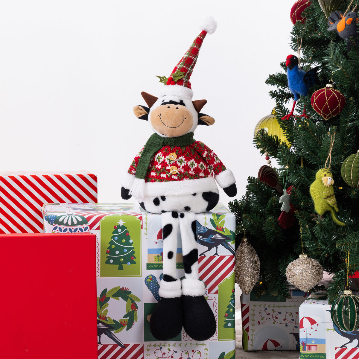 A Christmas-themed cow plush figurine wearing a red patterned sweater and a green-and-red Santa hat, posed on top of wrapped gift boxes beside a decorated Christmas tree.