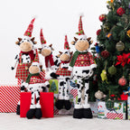 A set of Christmas cow plush figurines in different sizes, wearing festive red and green holiday sweaters, plaid skirts, and pointed red hats, posed next to a decorated Christmas tree and wrapped gifts.