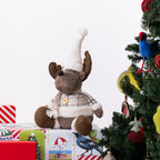 A plush moose figurine wearing a beige knit Santa hat, striped scarf, and plaid sweater, sitting on wrapped Christmas presents beside a decorated Christmas tree with a blue bird ornament.