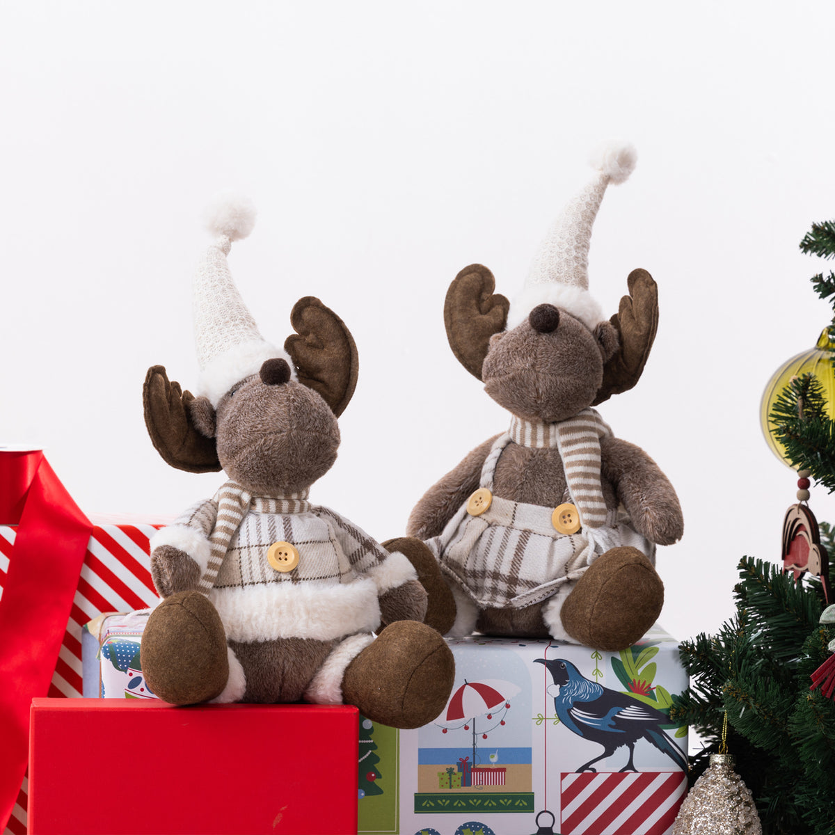 Two plush moose figurines dressed in holiday clothing, seated on top of wrapped Christmas presents with a tree and ornaments partially visible at the right edge, against a clean white background.