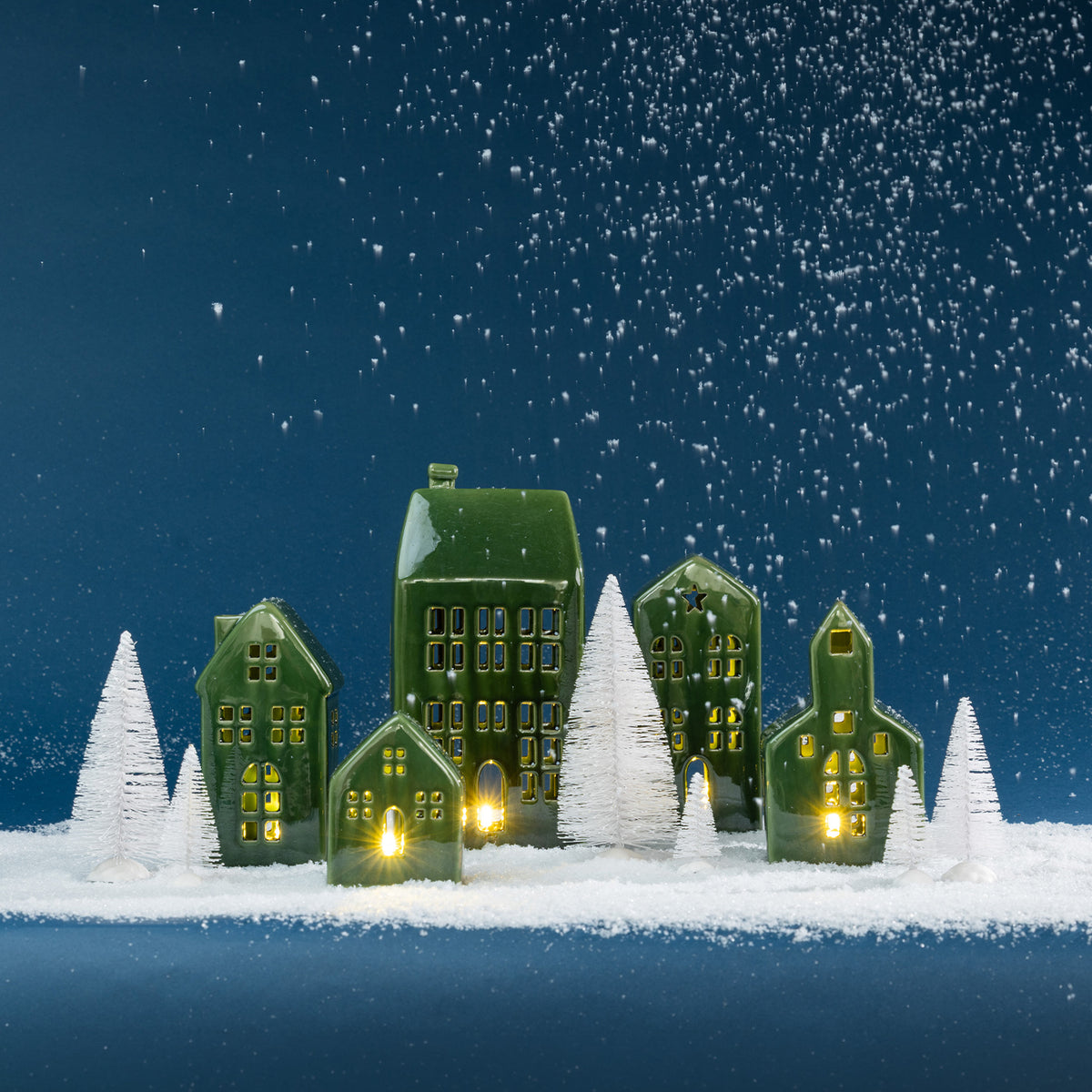 Christmas Wishes green ceramic village set illuminated among white snowy Christmas trees against a deep blue backdrop.