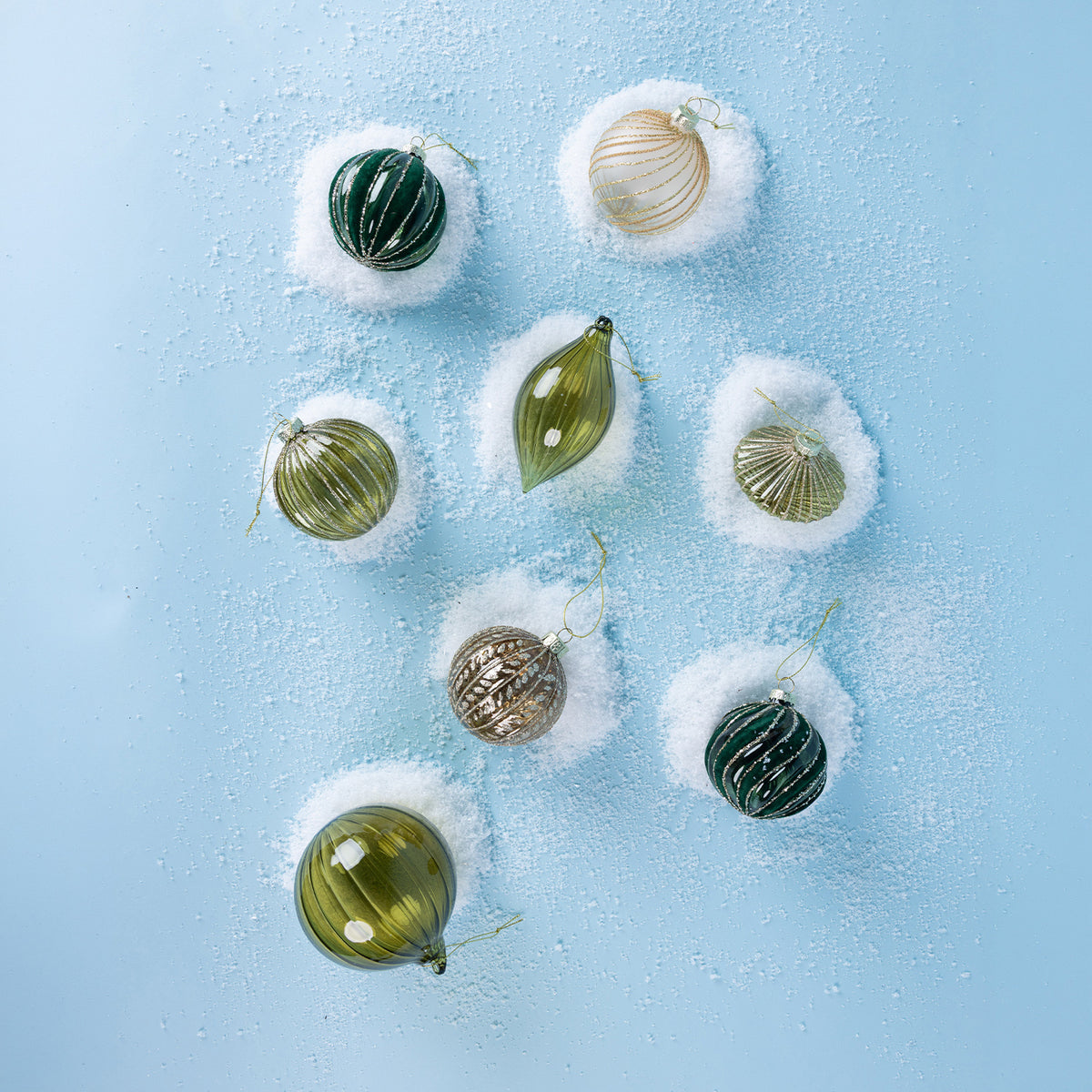 A collection of green, champagne and bronze glass Christmas baubles featuring glittered swirl and ribbed designs, displayed on a snowy light blue background.