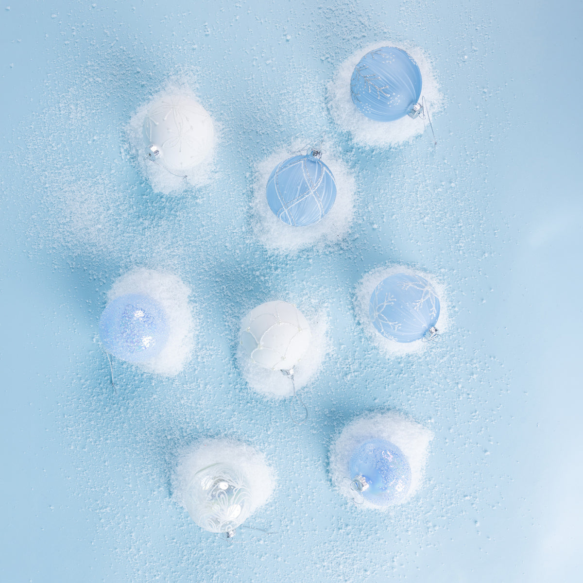 A collection of icy blue and white Christmas baubles featuring glittered snowflake, floral and scalloped designs, arranged on a snowy light blue background.