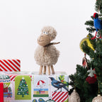 A plush sheep figurine with curly cream wool, legs, and a patterned scarf stands on top of wrapped Christmas gift boxes beside a decorated tree with a blue bird ornament.