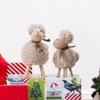 Two plush sheep figurines with curly beige wool, legs, and patterned scarves, posed on top of colorful gift boxes against a clean white background.