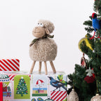 A plush sheep figurine with curly cream wool, legs, and a patterned scarf stands on top of wrapped Christmas gift boxes beside a decorated tree with a blue bird ornament.