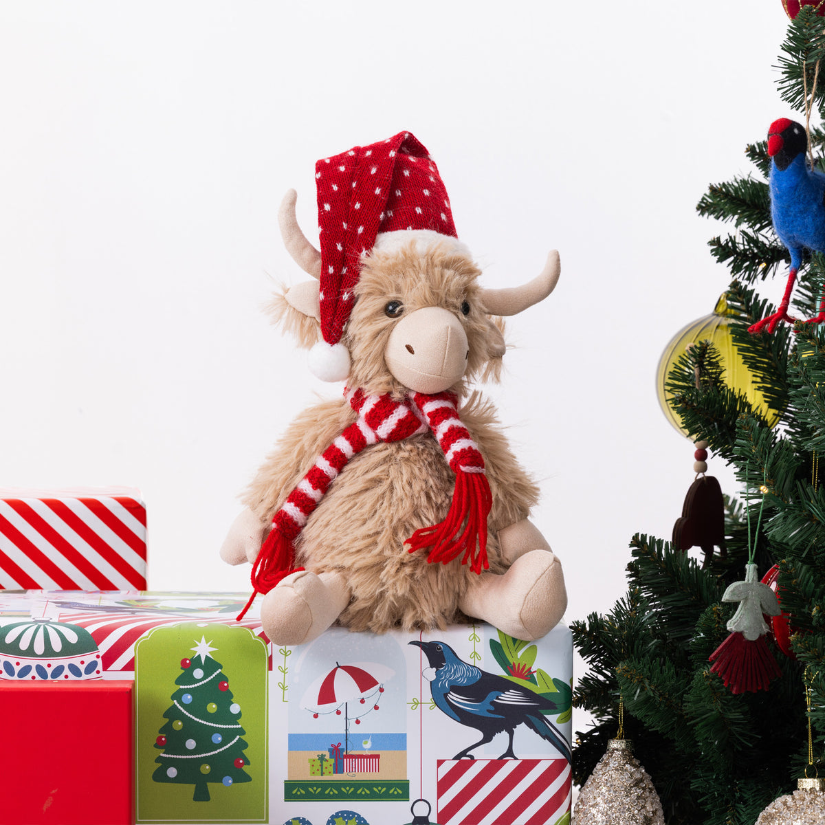 A fluffy Highland cow plush figurine wearing a red Santa hat and red-and-white striped scarf, sitting on top of wrapped Christmas presents beside a decorated tree.