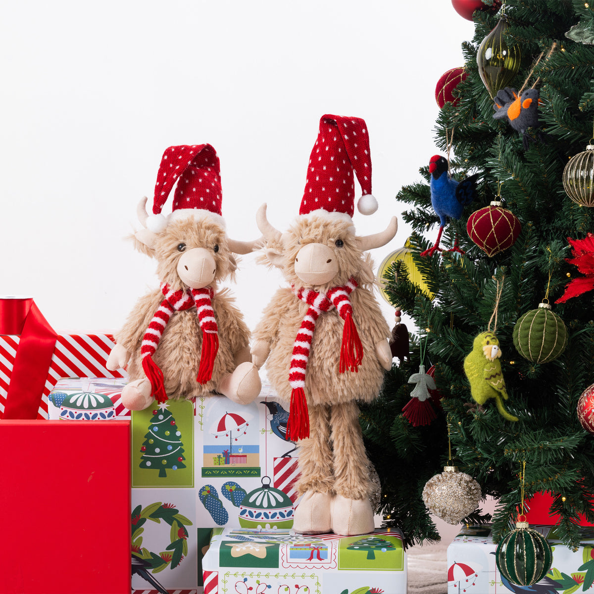 Two plush Highland cow figurines wearing red Santa hats and red-and-white striped scarves, standing on wrapped Christmas presents beside a decorated tree.