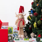 A fluffy Highland cow plush figurine wearing a red Santa hat and red-and-white striped scarf, standing on top of wrapped Christmas presents beside a decorated tree.