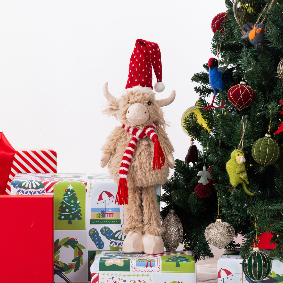 A fluffy Highland cow plush figurine wearing a red Santa hat and red-and-white striped scarf, standing on top of wrapped Christmas presents beside a decorated tree.