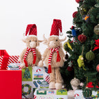 Two plush Highland cow figurines wearing red Santa hats and red-and-white striped scarves, standing on wrapped Christmas presents beside a decorated tree.