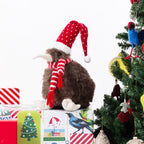 A plush kiwi bird figurine wearing a red Santa hat with white polka dots and a matching red-and-white striped scarf, sitting on top of wrapped Christmas presents beside a decorated Christmas tree on a white background.