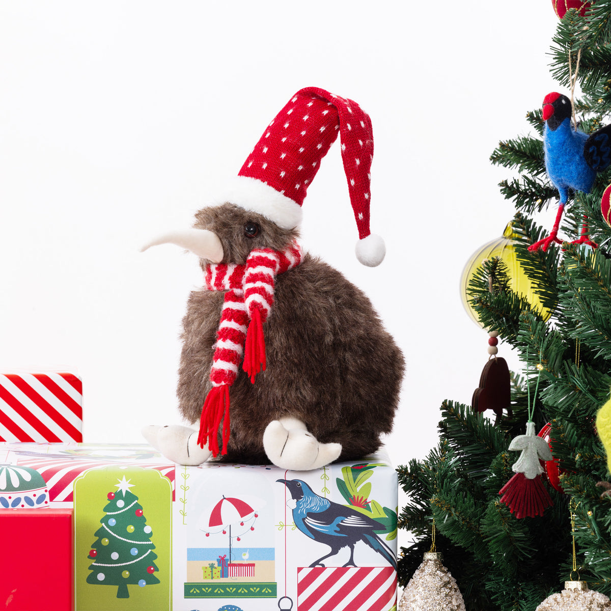 A plush kiwi bird figurine wearing a red Santa hat with white polka dots and a matching red-and-white striped scarf, sitting on top of wrapped Christmas presents beside a decorated Christmas tree on a white background.